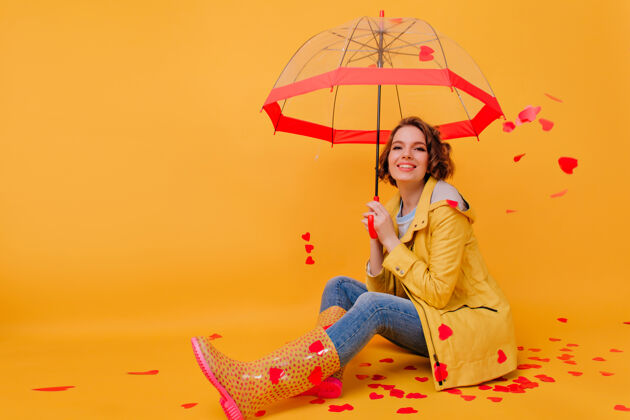 伞一位身穿黄色外套 面带微笑的年轻女子在墙上摆出红心的姿势美丽的女孩手持阳伞庆祝情人节雨魅力漂亮
