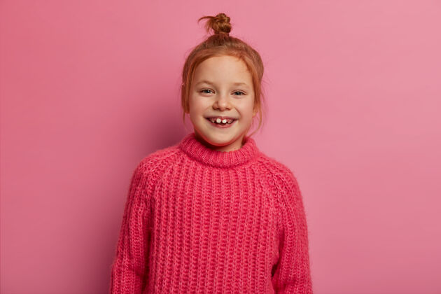 狐狸可爱的五岁女孩摆姿势 表达积极的情绪 有姜黄色的头发 穿着温暖的冬季毛衣 很高兴被拍照 对着粉色的墙壁摆姿势真诚的情感和孩子们乐观爱孩子