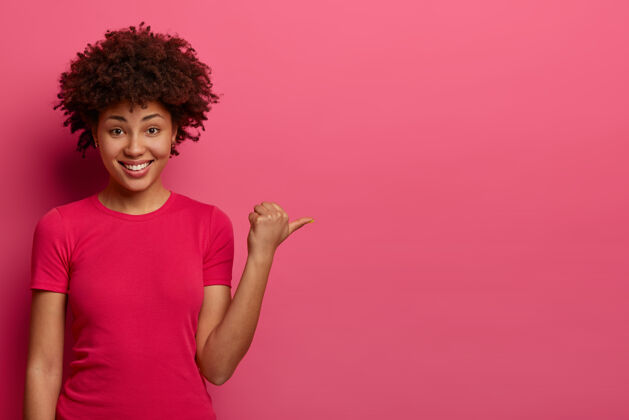 指向积极的年轻女店员帮助顾客找到更衣室 推销产品 把拇指放在一边 穿着休闲t恤 高兴地微笑 孤立在粉红色的墙上离开美国位置
