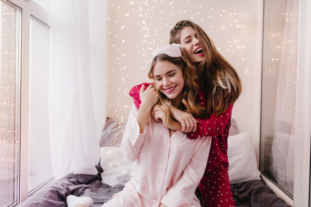 朋友穿着粉红色睡衣的快乐女孩坐在床上 眼睛闭着室内照片是快乐的欧洲女人微笑着拥抱她的妹妹睡衣能量友谊