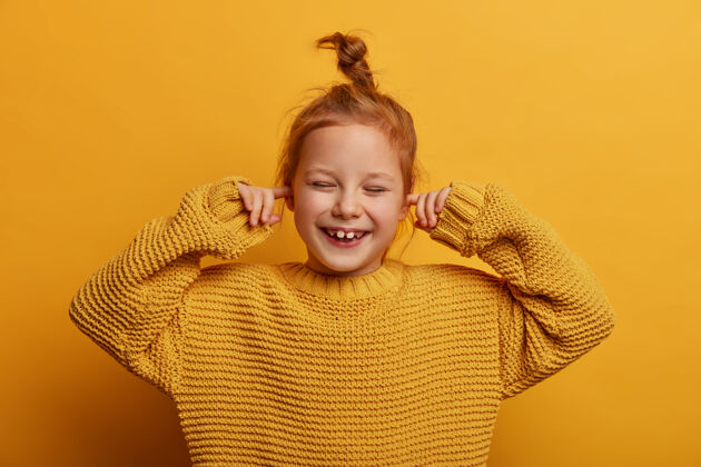 毛衣乐观快乐的小朋友的横拍用食指塞住耳朵 积极地笑 有姜黄色的发髻 穿着大号的针织毛衣 隔离在黄色的墙上停止这声音乐观满意结