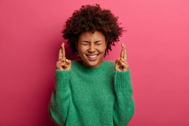 年轻迷信的非裔美国妇女专注于胜利 希望得到积极的结果 高兴地交叉手指和微笑 穿着绿色毛衣 预测重要的消息 孤立在粉红色的墙上卷发欢呼请