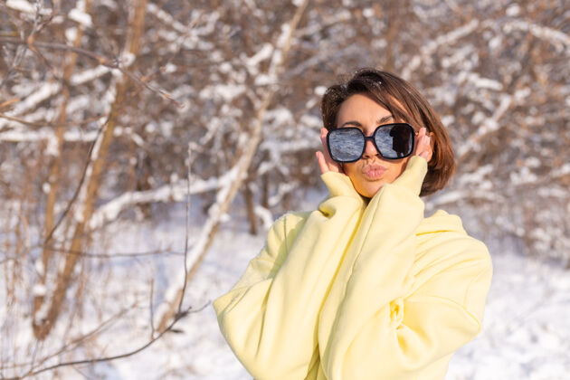 太阳阳光明媚的日子里 雪景冬林中一位年轻美女的画像 身着黄色大套头衫 戴着墨镜 享受着阳光和雪美丽女人白牙