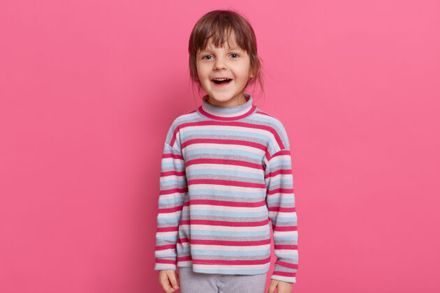 庆祝快乐兴奋的学龄前女孩穿着休闲风格的条纹衬衫在粉红色的墙上摆出孤立的姿势深色头发站立兴奋