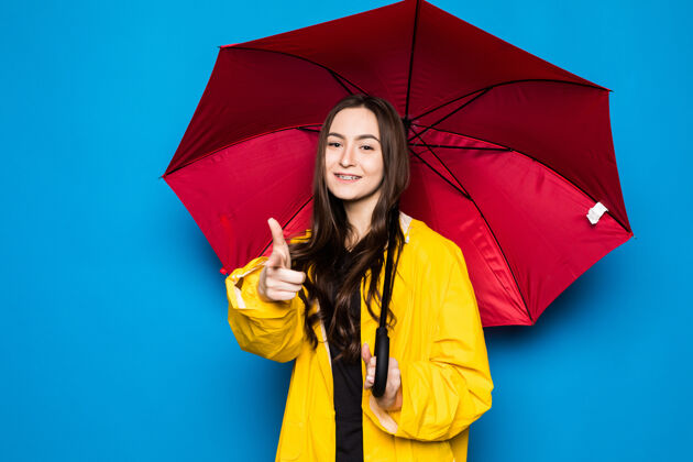 天快乐的年轻女子拿着黄色雨衣和蓝色墙壁的雨伞成功模特表情