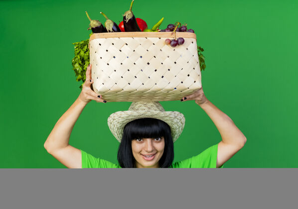 女高兴的年轻女园丁穿着制服 戴着园艺帽 把菜篮子举过头顶篮子蔬菜绿色