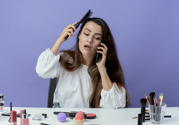 化妆震惊的美丽女孩坐在桌子旁用化妆工具梳头讲电话隔离在紫色的墙上头发桌子美女
