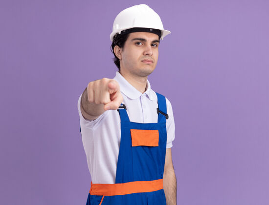 建筑年轻的建筑工人穿着建筑制服 戴着安全帽 食指指着前面 站在紫色的墙壁上不高兴地看着建筑工人男人目录
