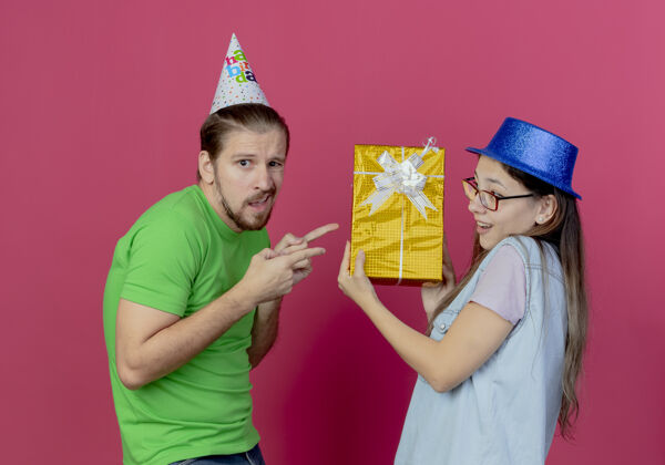 粉色一个戴着派对帽的年轻人指着挂在粉红色墙上的戴着蓝色派对帽的女孩手里的礼盒礼物指着聚会