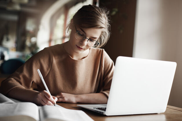 企业家戴着时髦眼镜的勤劳专注的女人专注于写文章 坐在笔记本电脑旁舒适的咖啡馆里 认真地工作和做笔记室内信息互联网