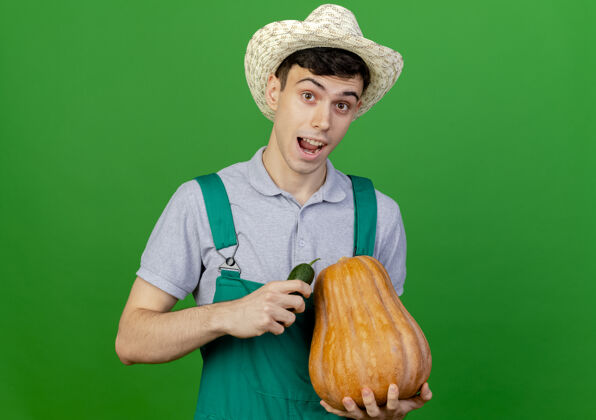 穿快乐的年轻男园丁戴着园艺帽 手里拿着黄瓜和南瓜花园绿色黄瓜
