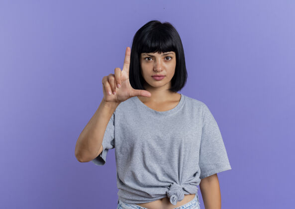 枪自信的年轻黑发白种女人手势标志孤立的紫色背景与复制空间自信背景手