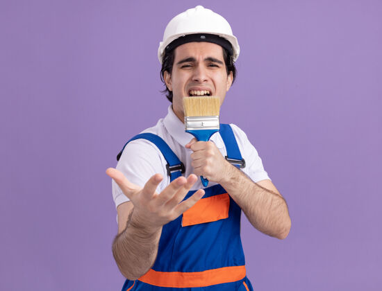 男人有趣而快乐的年轻建筑工人穿着建筑制服 戴着安全帽 拿着油漆刷 拿着麦克风 站在紫色的墙上唱歌麦克风制服欢乐