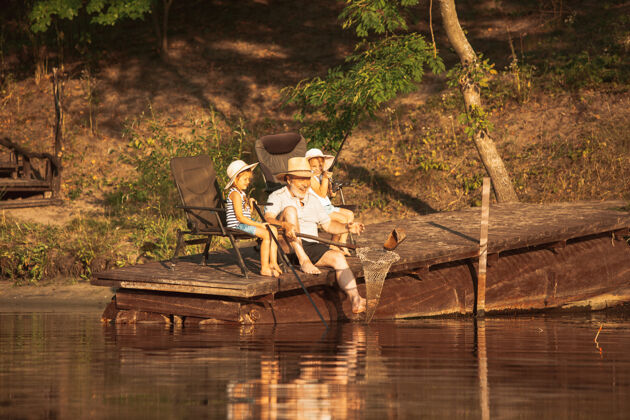 兴奋可爱的小女孩和他们的爷爷在湖边或河边钓鱼在夏天的日落时分 在靠近水和森林的码头上休息家庭 娱乐 童年 自然的概念坐着自然活动