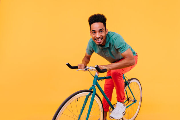 年轻穿着绿色t恤和白色运动鞋的时髦非洲男人在自行车上摆姿势骑在自行车上的幸福的黑人男人明亮非洲活动