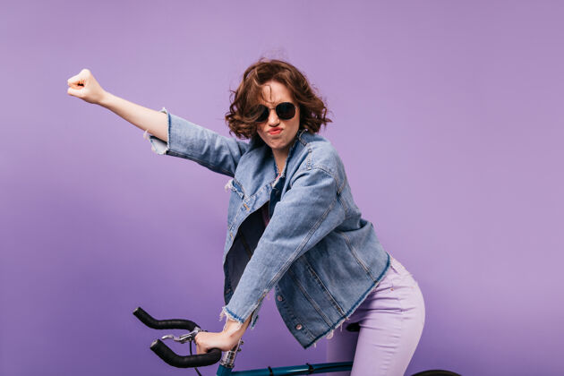 寒冷穿着休闲服的有趣女孩在自行车上摆姿势无忧无虑的卷发女士坐在自行车上的肖像肖像乐趣休闲
