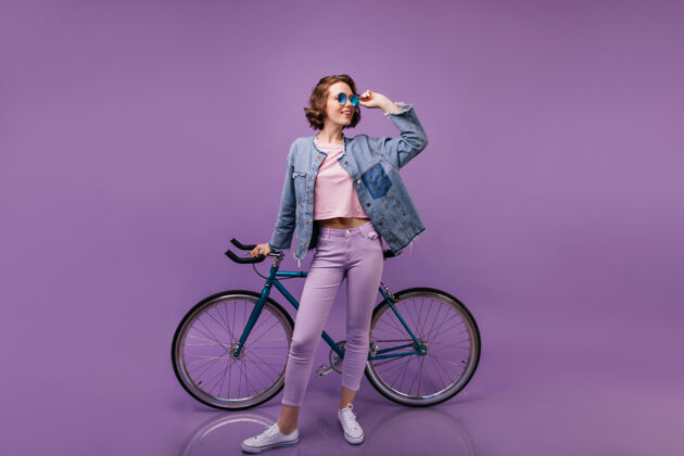 运动员苗条活泼的女人与自行车合影紫色裤子的卷曲女模特的室内全长镜头女士快乐微笑
