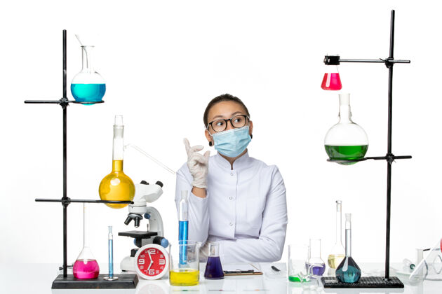 坐前视图穿着医疗服的女化学家 戴着面罩坐在桌子前 溶液背景为浅白色病毒化学实验室的covidsplash桌子设备西装