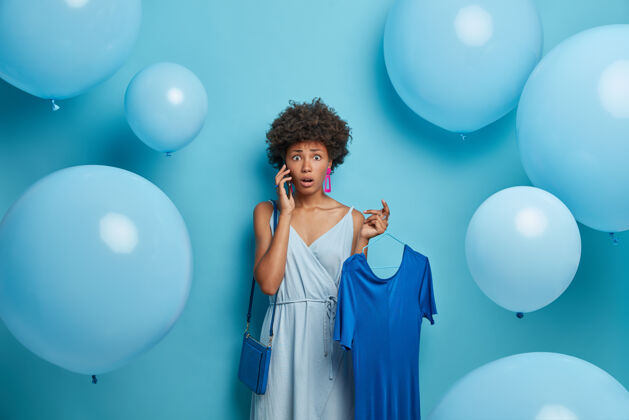 情感震惊的女人通过手机和朋友聊天 发现了令人兴奋的消息 喜欢蓝色 把衣服挂在衣架上 穿着出门的衣服 站在室内用氦气球 有着困惑和担心的神情惊喜室内手机