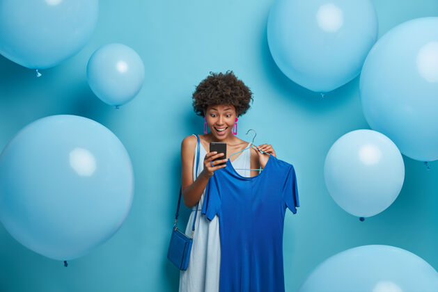 装置生日 节日 服装概念令人印象深刻的快乐女人用惊奇的快乐目光盯着智能手机显示屏 收到意想不到的信息 在衣架上挑选衣服 穿上所有蓝色的衣服优雅衣架选择