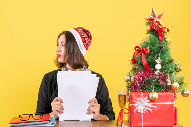 圣诞老人穿着西装 戴着圣诞老人帽和新年装饰品 手里拿着文件 坐在办公桌旁 桌上放着圣诞树坐着年文件