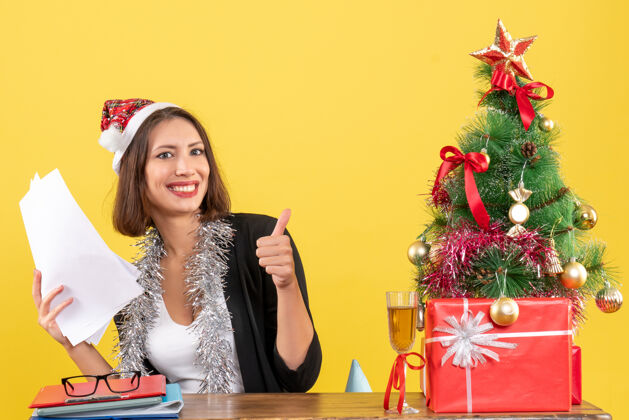文件穿着西装 戴着圣诞老人帽 戴着新年装饰品 拿着文件 做着“ok”的手势 坐在一张桌子旁 桌上放着圣诞树树西装坐着