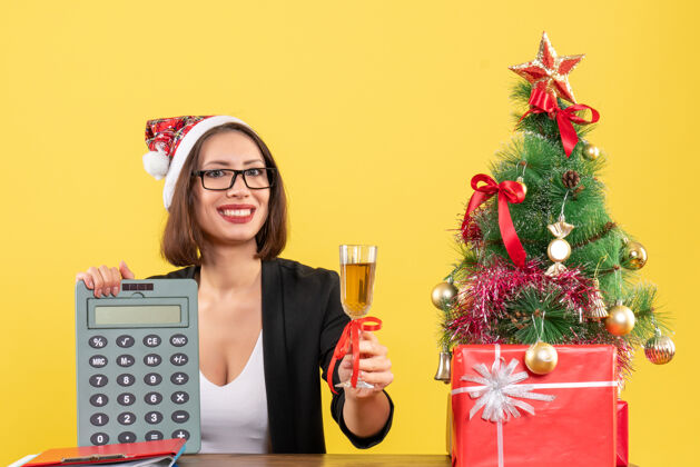 帽子微笑迷人的女士穿着西装 戴着圣诞老人的帽子和眼镜 在黄色的办公室里展示计算器和举酒圣诞老人提高模特