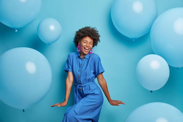 心情无忧无虑快乐的卷发女人快乐地跳舞 穿着蓝色的连衣裙 在派对上围着充气的氦气球发冷 感觉好玩 享受喜爱的节日 有乐观的节日心情快乐的时刻气球寒冷氦