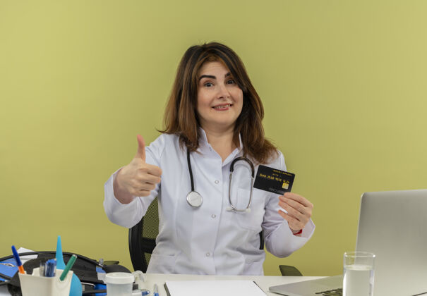 拇指微笑的中年女医生穿着医用长袍和听诊器坐在办公桌旁 手里拿着医疗工具和手提电脑 手里拿着信用卡 大拇指向上伸出向上显示个人