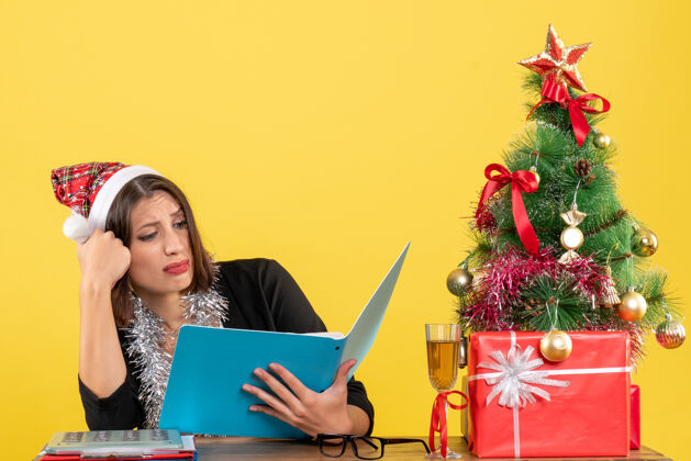微笑穿着西装 戴着圣诞老人帽和新年装饰品的商务女士专注于文件 坐在办公室的桌子旁 桌上放着圣诞树电脑文件圣诞老人