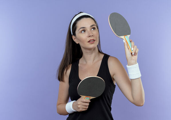 站立戴着头巾拿着球拍打乒乓球的年轻健身女士站在蓝色背景上 自信地看着一边持球头带年轻
