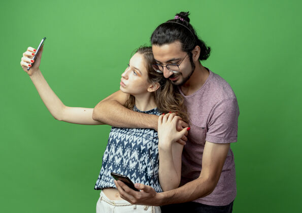 同时一对年轻的男女 幸福的男人拥抱着他的女朋友 而她用智能手机拍摄他们站在绿色背景下的照片情侣拍照拥抱
