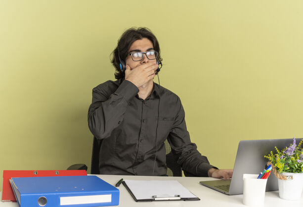 坐着年轻震惊的上班族戴着耳机戴着光学眼镜坐在办公桌旁使用办公工具 看着笔记本电脑把手放在嘴上 隔离在绿色背景上 留有复印空间工具眼镜年轻