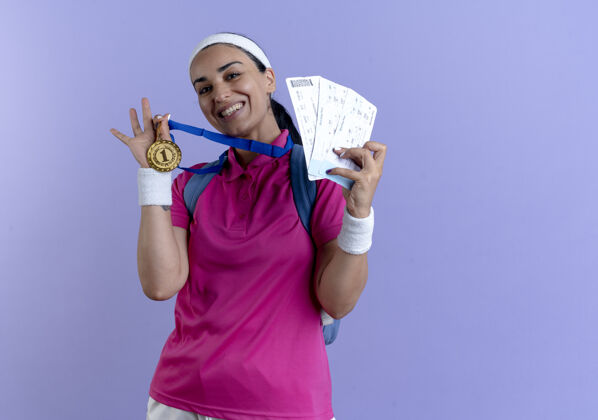 门票年轻的笑容可掬的白人运动女性 戴着背包头带和腕带 手持金牌和机票 背景为紫色 留有复印空间背部包年轻
