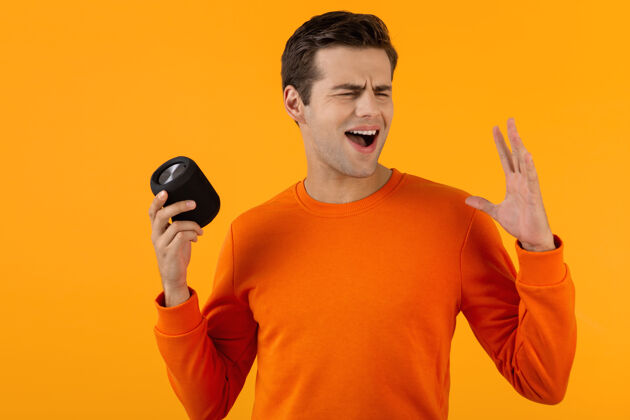 室内时尚微笑的年轻人穿着橙色毛衣手持无线扬声器快乐地听音乐设备姿势便携