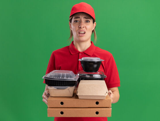 感觉穿着制服的年轻漂亮的送货员拿着纸食品包装和比萨盒上的容器 这些比萨盒被隔离在绿色的墙上姿势制服年轻人