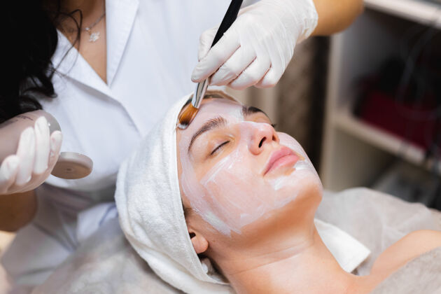 年轻美容师用刷子将白色保湿面膜涂在spa美容院一位年轻女客户的脸上Spa治疗健康
