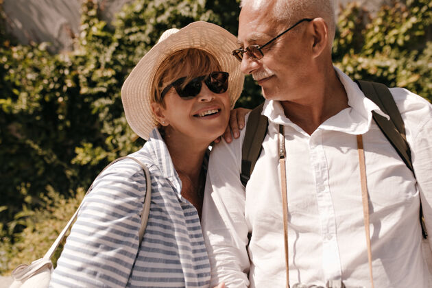 退休戴着太阳镜 蓝色衣服和帽子的积极的金发女士微笑着 在户外和穿着白衬衫的灰色头发男人合影城市一起老