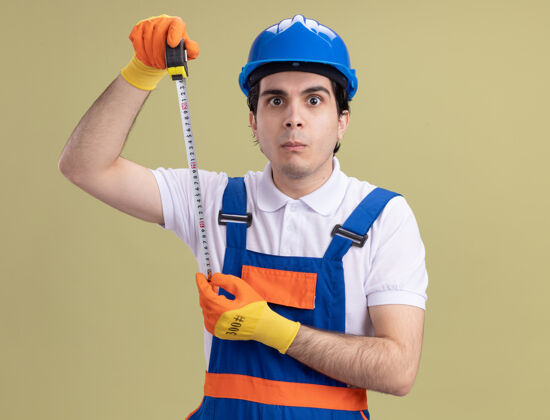 建筑工人年轻的建筑工人穿着建筑制服 戴着安全帽 戴着橡胶手套 手里拿着卷尺 站在绿色的墙壁上困惑地看着前方制服持有胶带