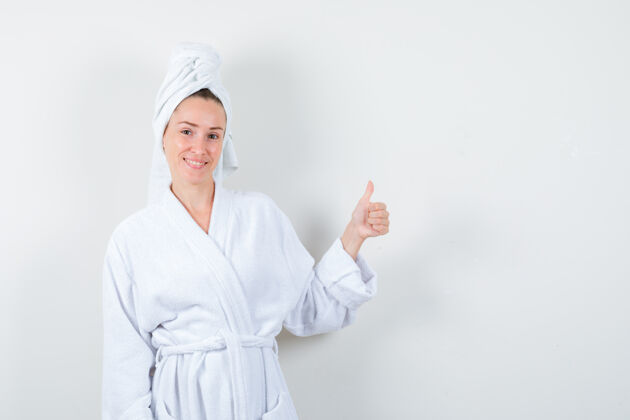 积极在白色浴袍 毛巾和看起来愉快的正面视图显示拇指向上的年轻女子的肖像乐趣女孩友谊
