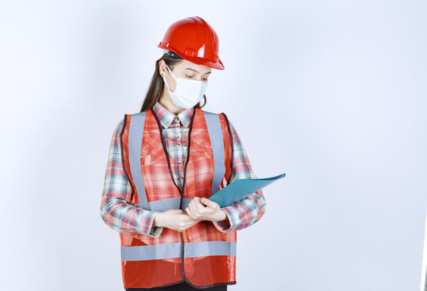 工作场所戴着安全面具 头戴红色头盔 手拿蓝色文件夹的女建筑工程师面罩人姿势