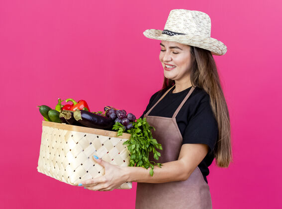微笑微笑着美丽的园丁女孩穿着制服 戴着园艺帽 拿着蔬菜篮子看着粉红色的背景举行花园帽子