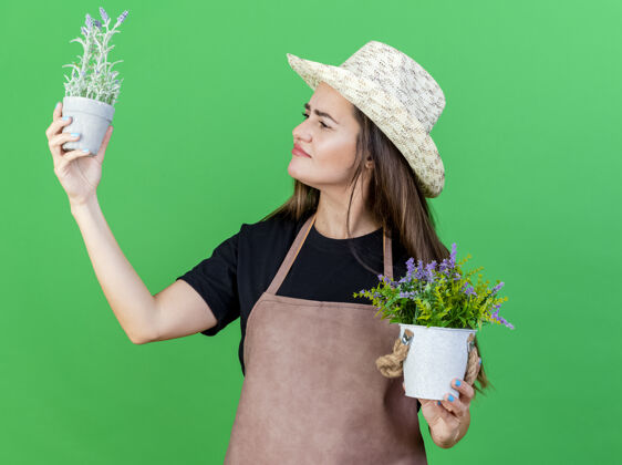 花盆高兴美丽的园丁女孩穿着制服 戴着园艺帽 抬头望着绿色背景上孤立的花盆里的花朵花女孩制服