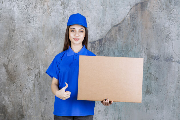 快递身着蓝色制服的女服务人员手持一个纸板箱 并显示积极的手势工人送货满意