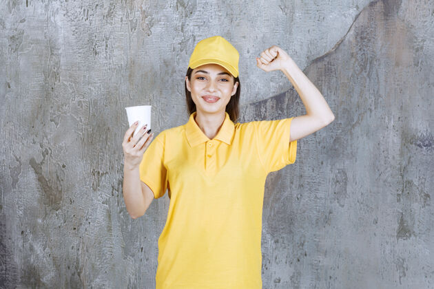 快递身着黄色制服的女服务人员手持塑料杯 并显示积极的手势享受交易年轻
