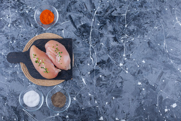 砧板鸡胸肉放在砧板上 放在三角架上 旁边是盛满香料的碗 放在蓝色的桌子上三脚架碗肉类