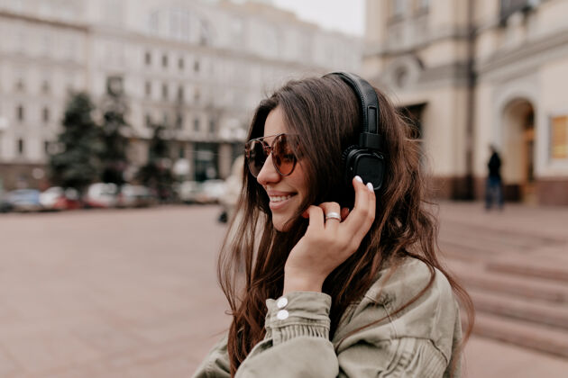 耳机一头黑发 穿着卡其色夹克 戴着时髦眼镜 戴着耳机听音乐 在老城街头漫步的兴奋快乐的女孩积极新潮欢乐