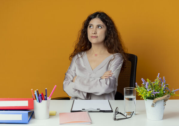 工人看着一边思考的年轻漂亮的女上班族坐在办公桌旁 办公工具交叉着双手 孤立地放在橙色的背景上年轻桌子漂亮