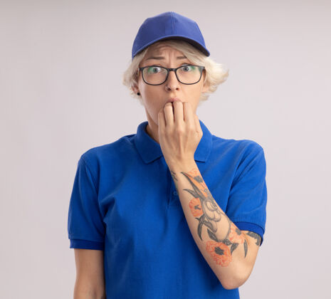 女人身穿蓝色制服 头戴鸭舌帽的年轻女送货员紧张地站在白色的墙上咬指甲站立分娩紧张