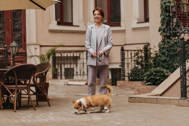 女人穿着灰色西装的女士带着她的高尔基犬散步穿着夹克和裤子的快乐女人和狗在外面合影的肖像城市咖啡馆女性